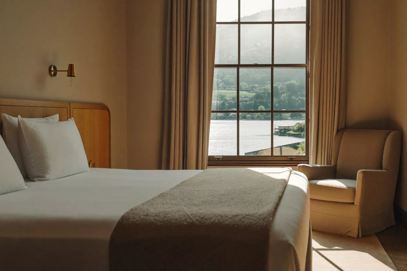 Chambre d'un hôtel indépendant romand, lit fait, fauteuil et grande fenêtre ouvrant sur le lac en fin de matinée